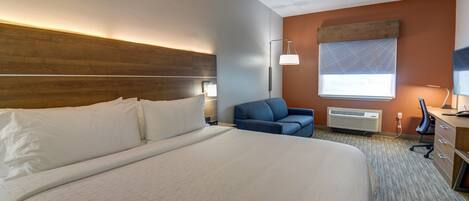 A hotel room with a large bed, a desk with a computer, a chair, a lamp, and a window with blinds.