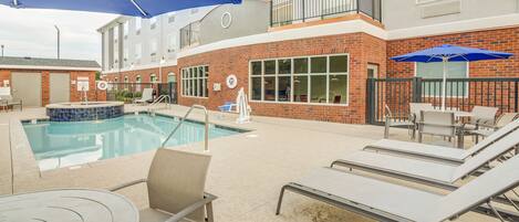 A pool area with lounge chairs and umbrellas, a brick building with large windows, and a small pool.