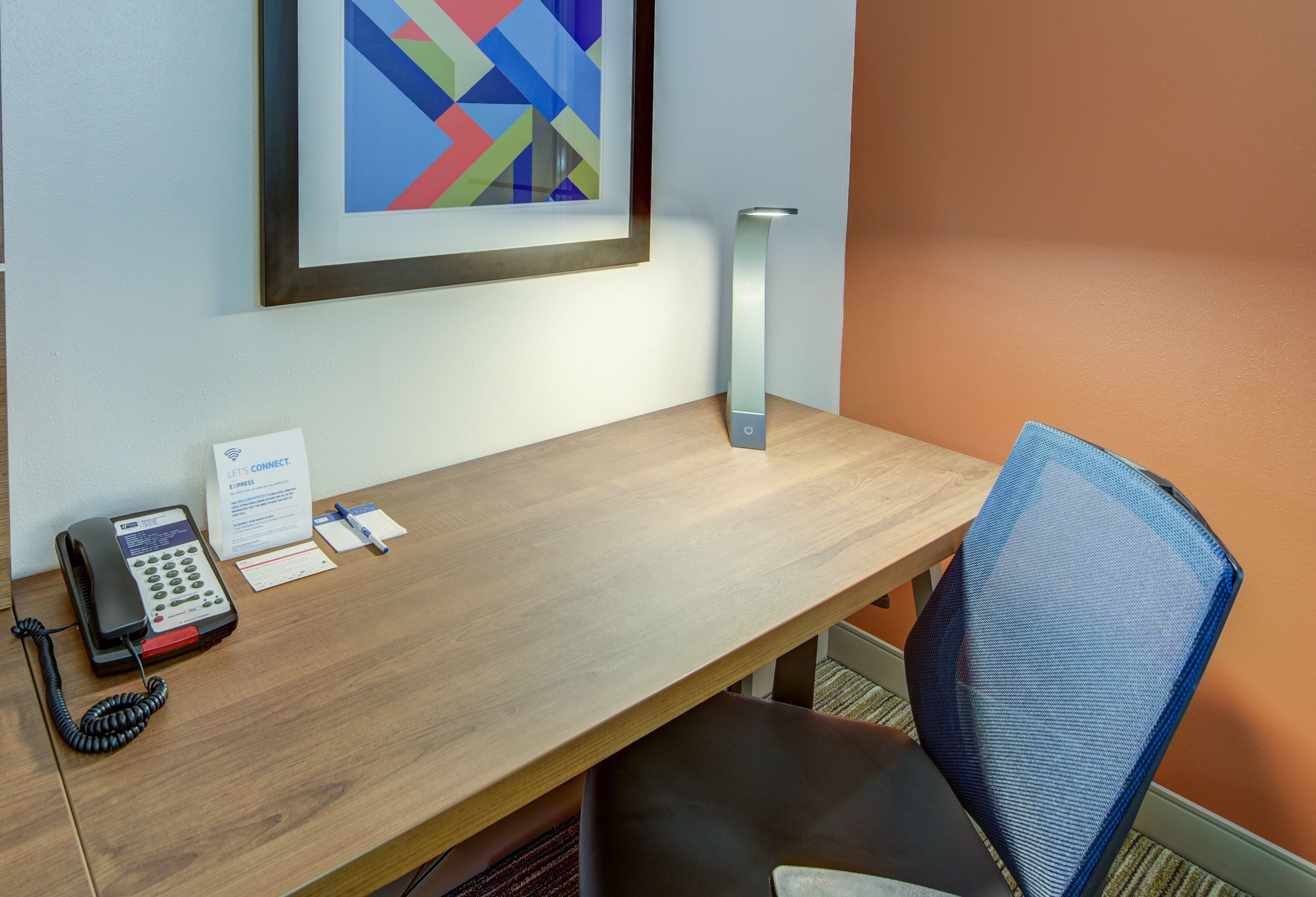 Een hotelkamer met een houten bureau, een telefoon, een lijst met abstract kunstwerk en een blauwe mesh-stoel.
