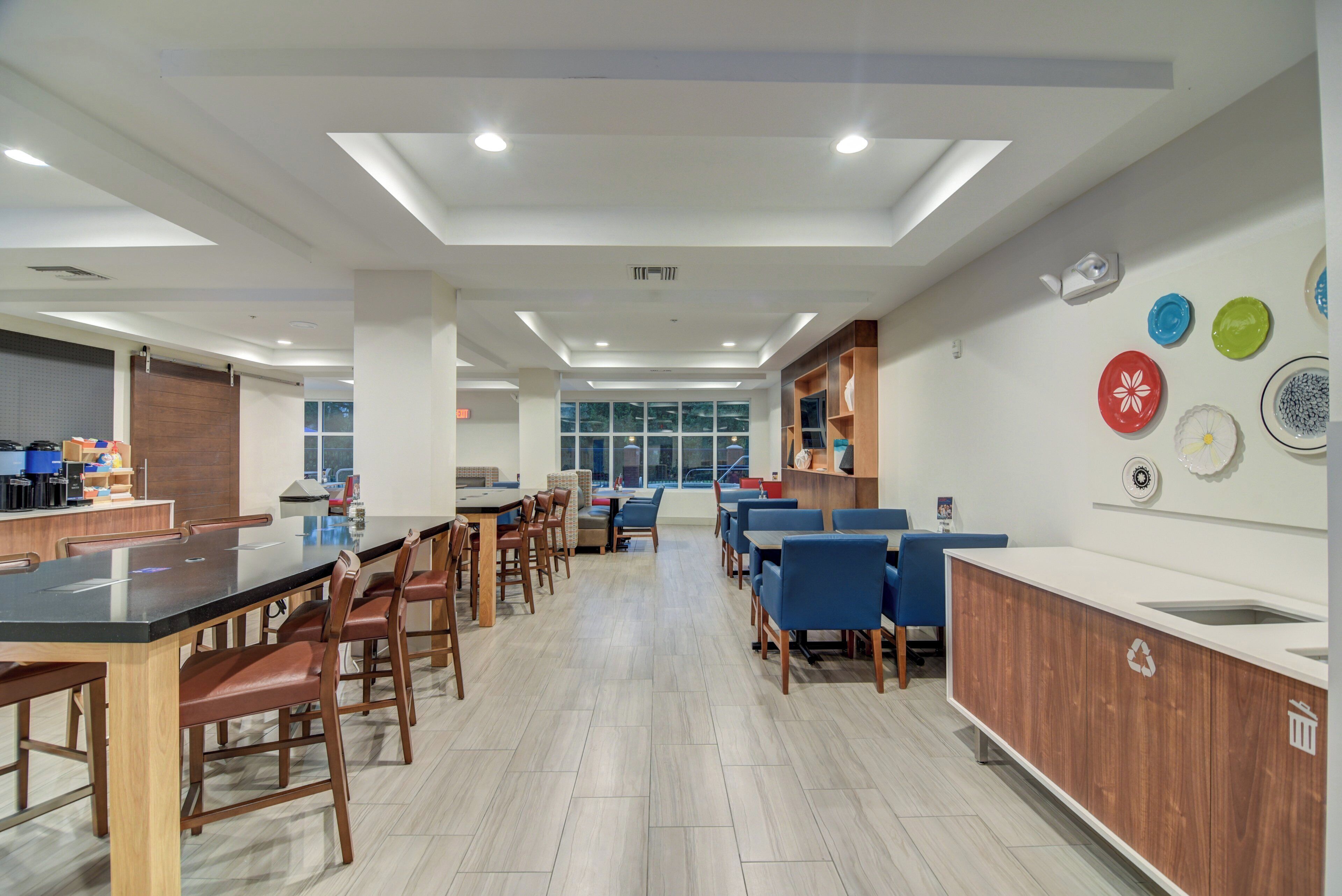A modern dining area with multiple tables and chairs, a kitchen counter, and a wall with decorative plates.