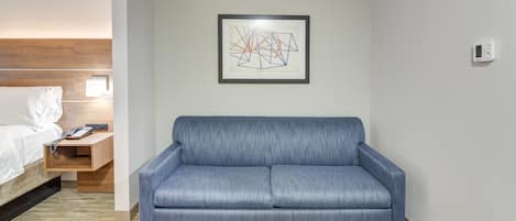 A modern hotel room with a blue sofa, a wooden bedside table, a white bed, and a framed abstract artwork on the wall.