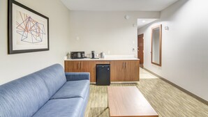 Moderni hotellihuone, jossa on sininen sohva, puinen sohvapöytä, pieni keittiö mikroaaltouunilla ja astianpesukoneella.