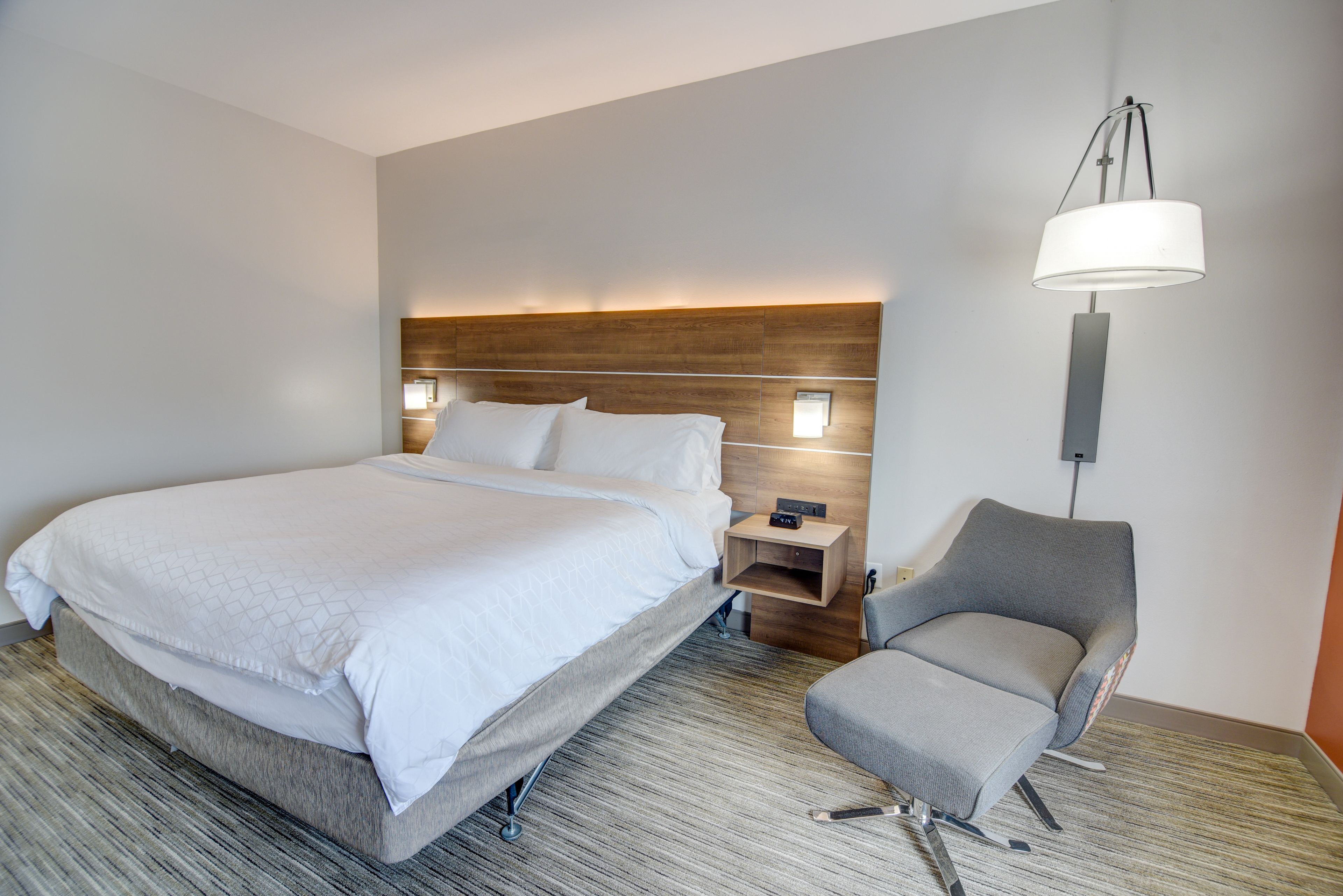 A hotel room with a large bed, a grey armchair, a bedside table, and a wall-mounted lamp.
