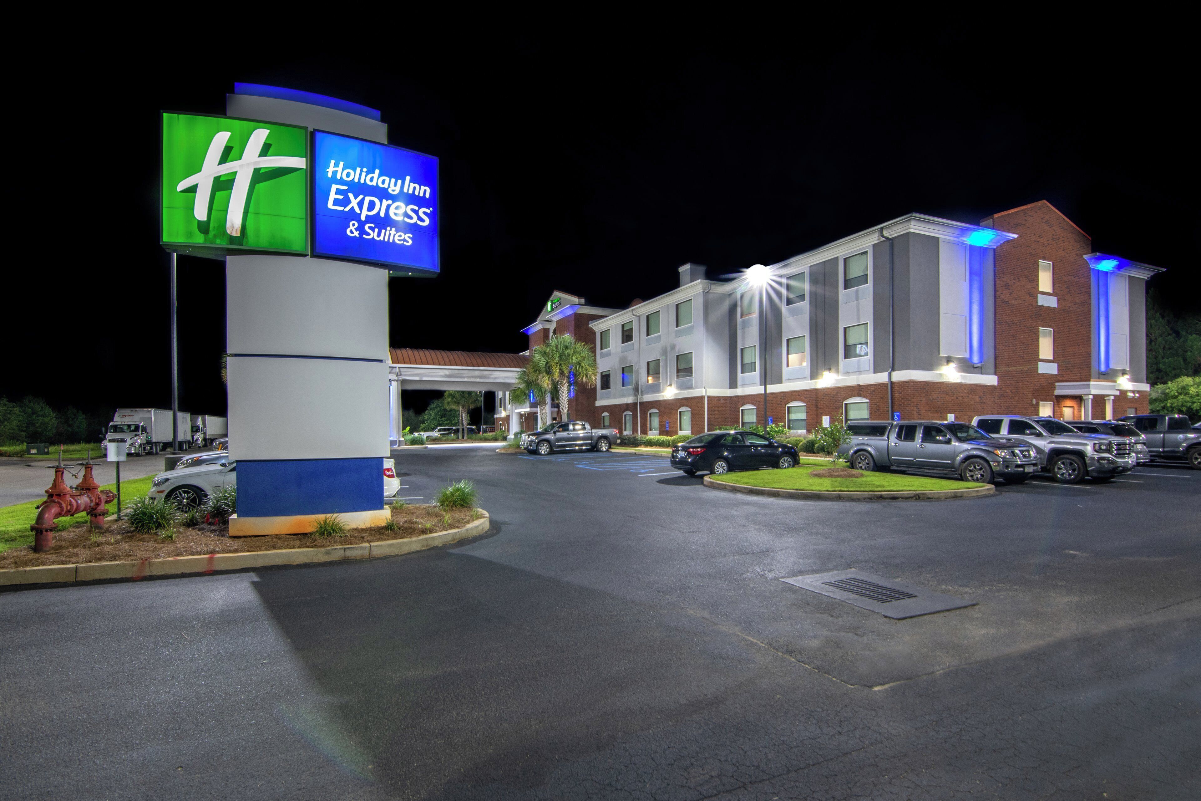 A Holiday Inn Express & Suites hotel with a large illuminated sign, a parking lot with several cars, and a building with multiple windows and a brick facade.