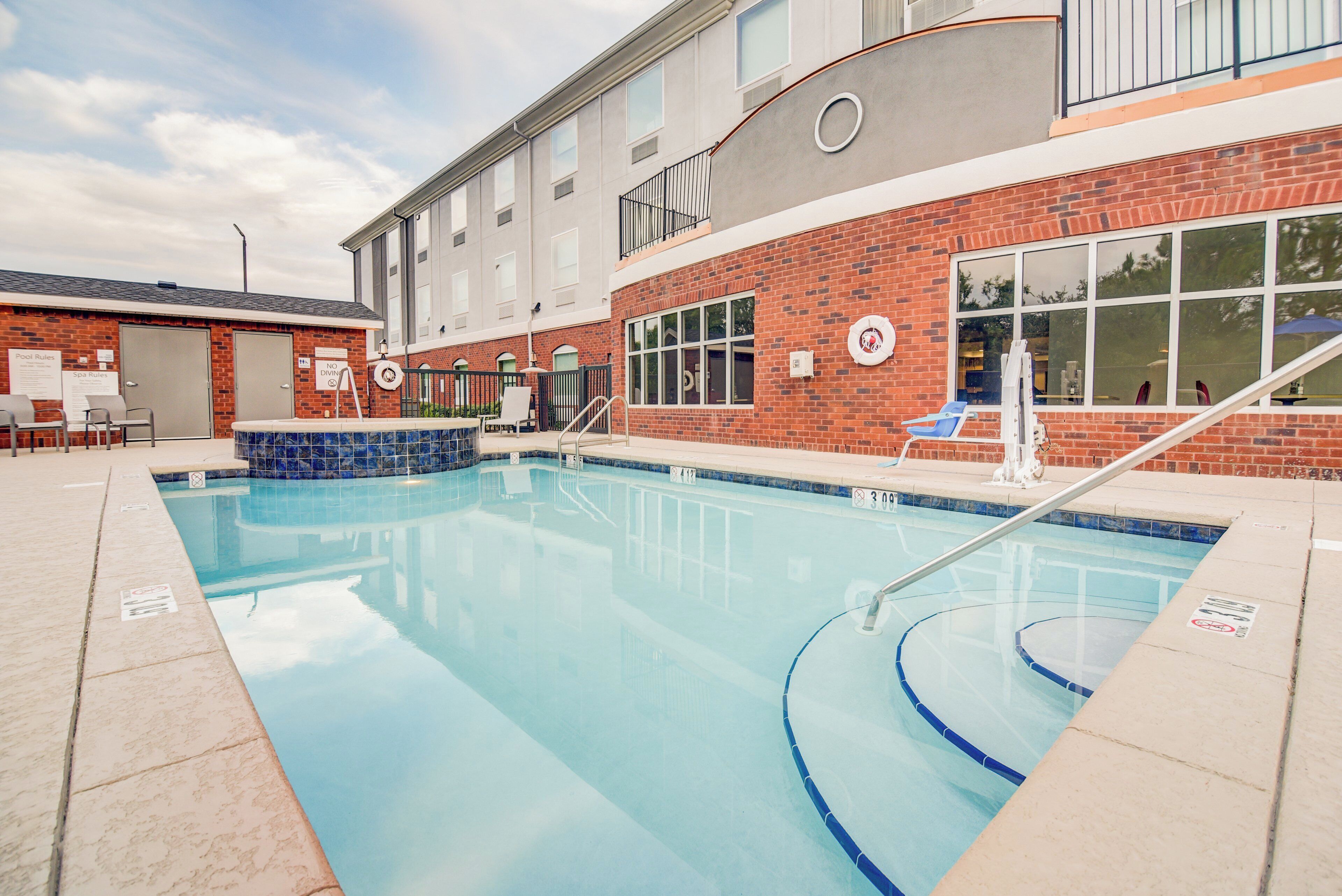 Une piscine à l’eau d’un bleu cristallin, entourée d’un bâtiment en briques doté de fenêtres et d’un espace couvert avec des chaises.
