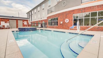 A swimming pool with a clear blue water, surrounded by a brick building with windows and a covered area with chairs.