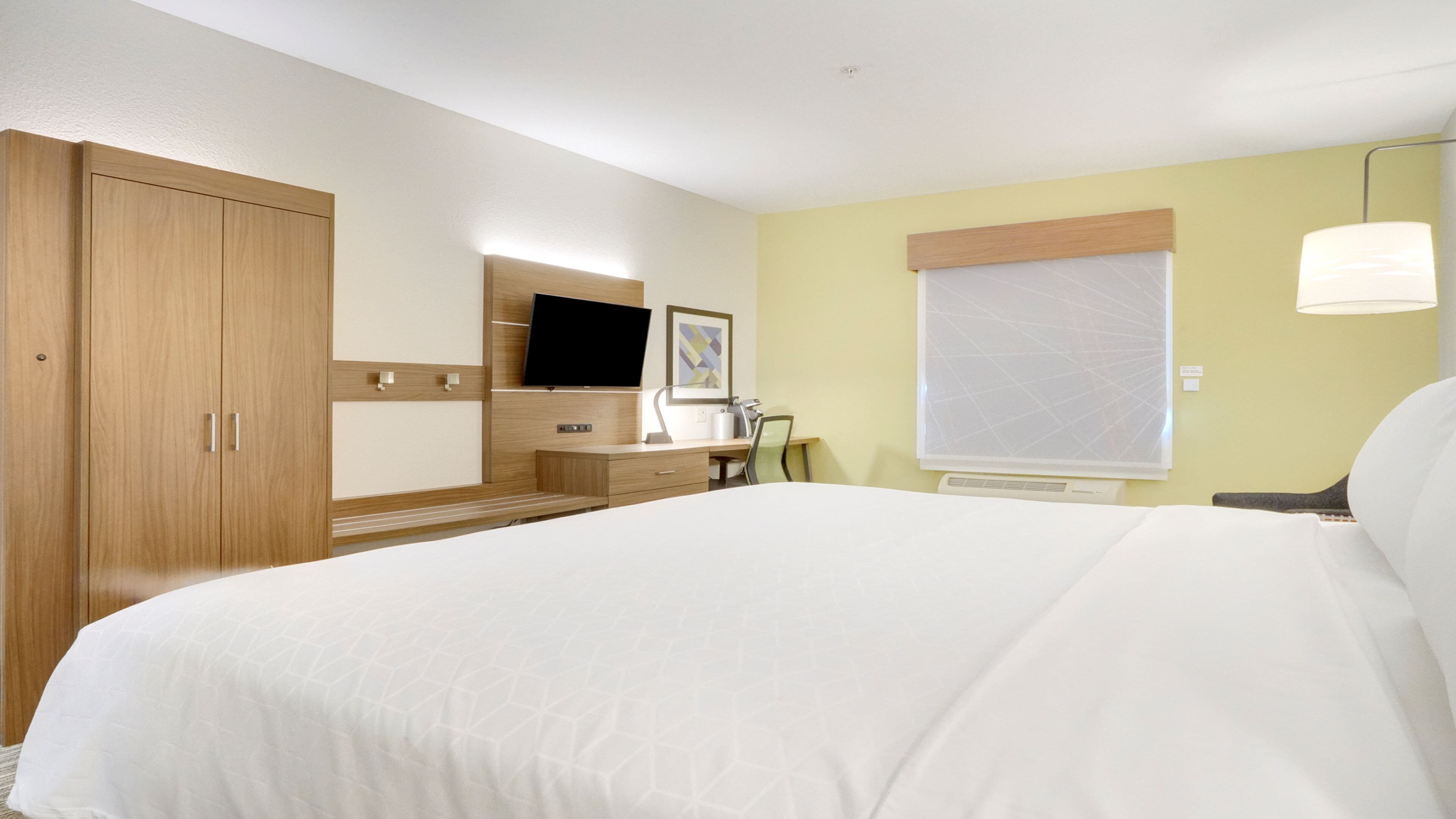 A hotel room with a large bed, a desk with a chair, a television, and a window with blinds.