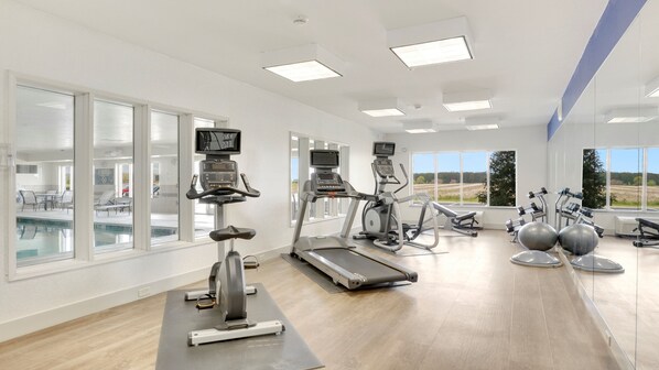 A gym with cardio machines, a pool, and large windows offering a view of the outdoors.