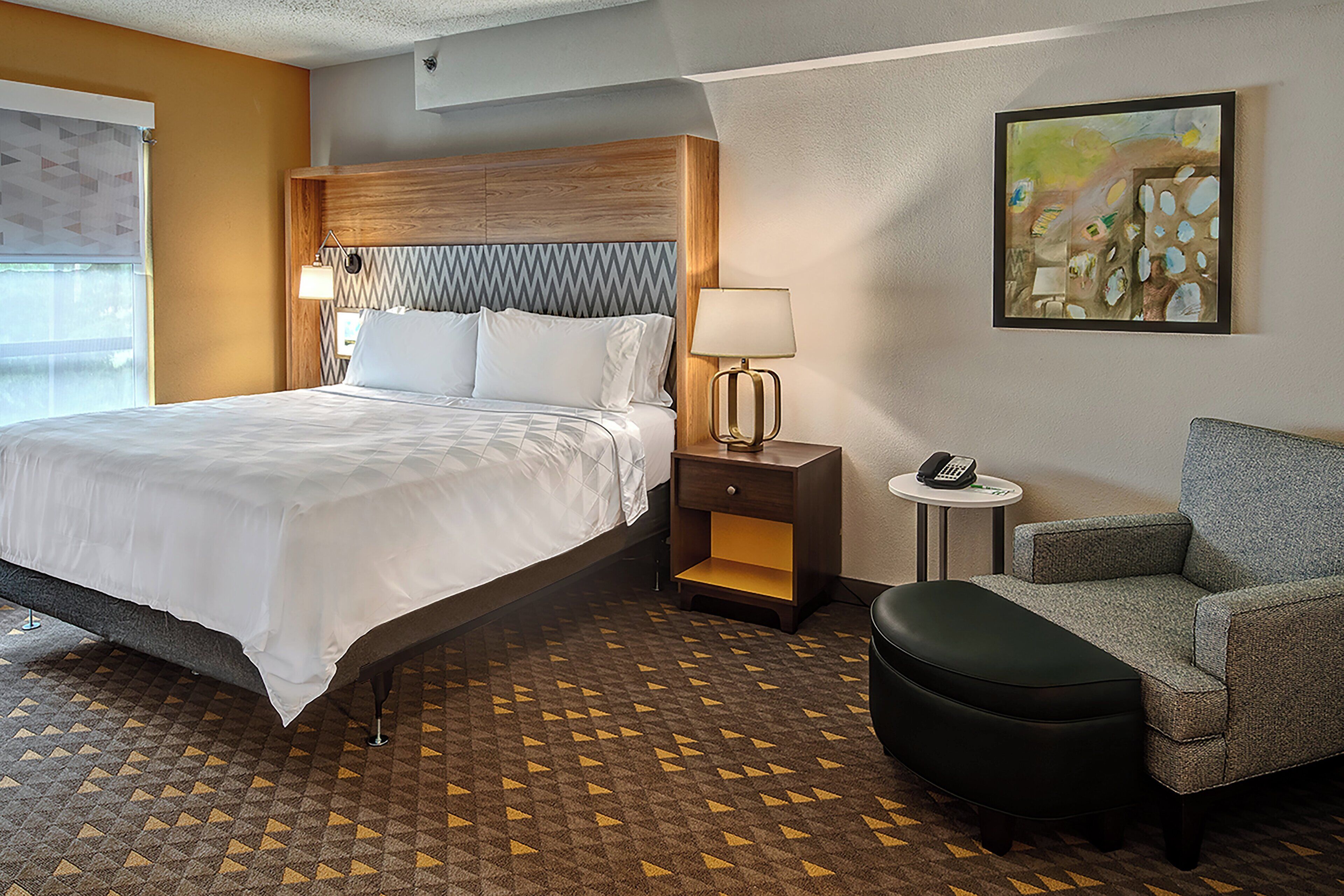 Una habitación de hotel con cama, mesita de noche, lámpara, silla y una obra de arte en la pared.