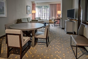 A hotel room with a round dining table, two chairs, a sofa, a TV, and a desk with a chair.