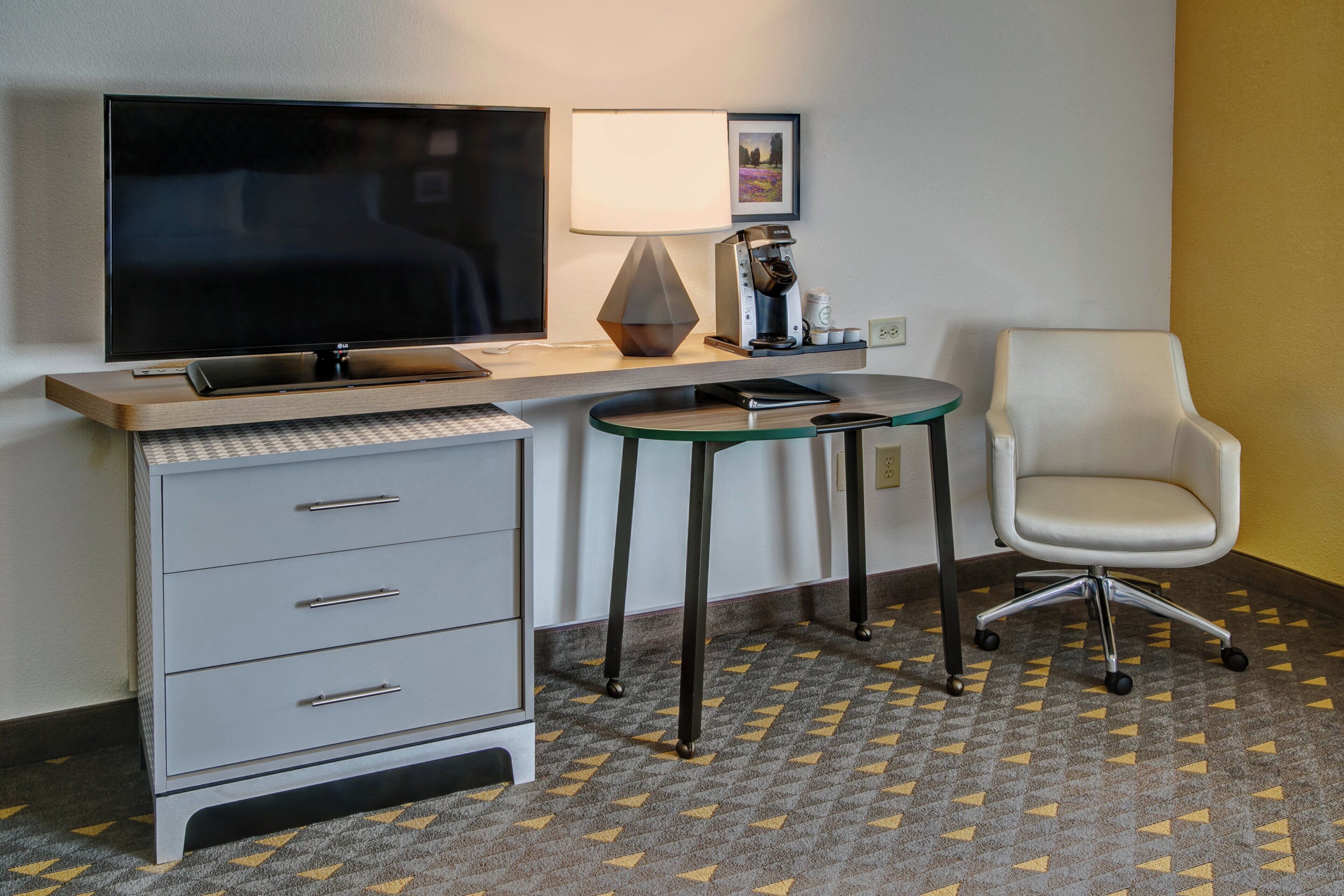 A hotel room with a desk, a flat-screen TV, a lamp, a chair, and a patterned carpet.