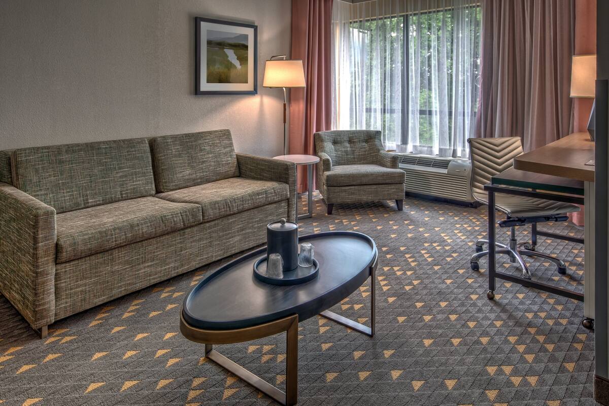 Une chambre d’hôtel avec un canapé, un fauteuil, une petite table avec une lampe et une fenêtre avec des rideaux.