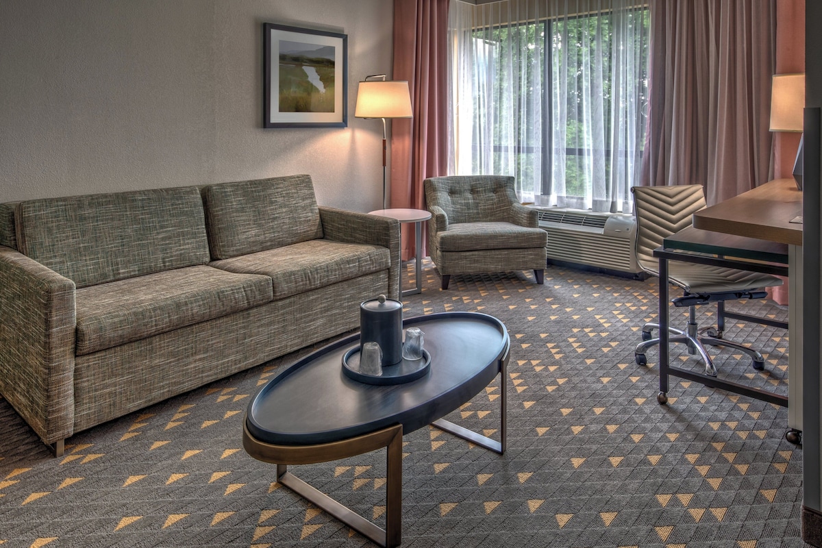 A hotel room with a sofa, armchair, small round table, and a desk with a chair.