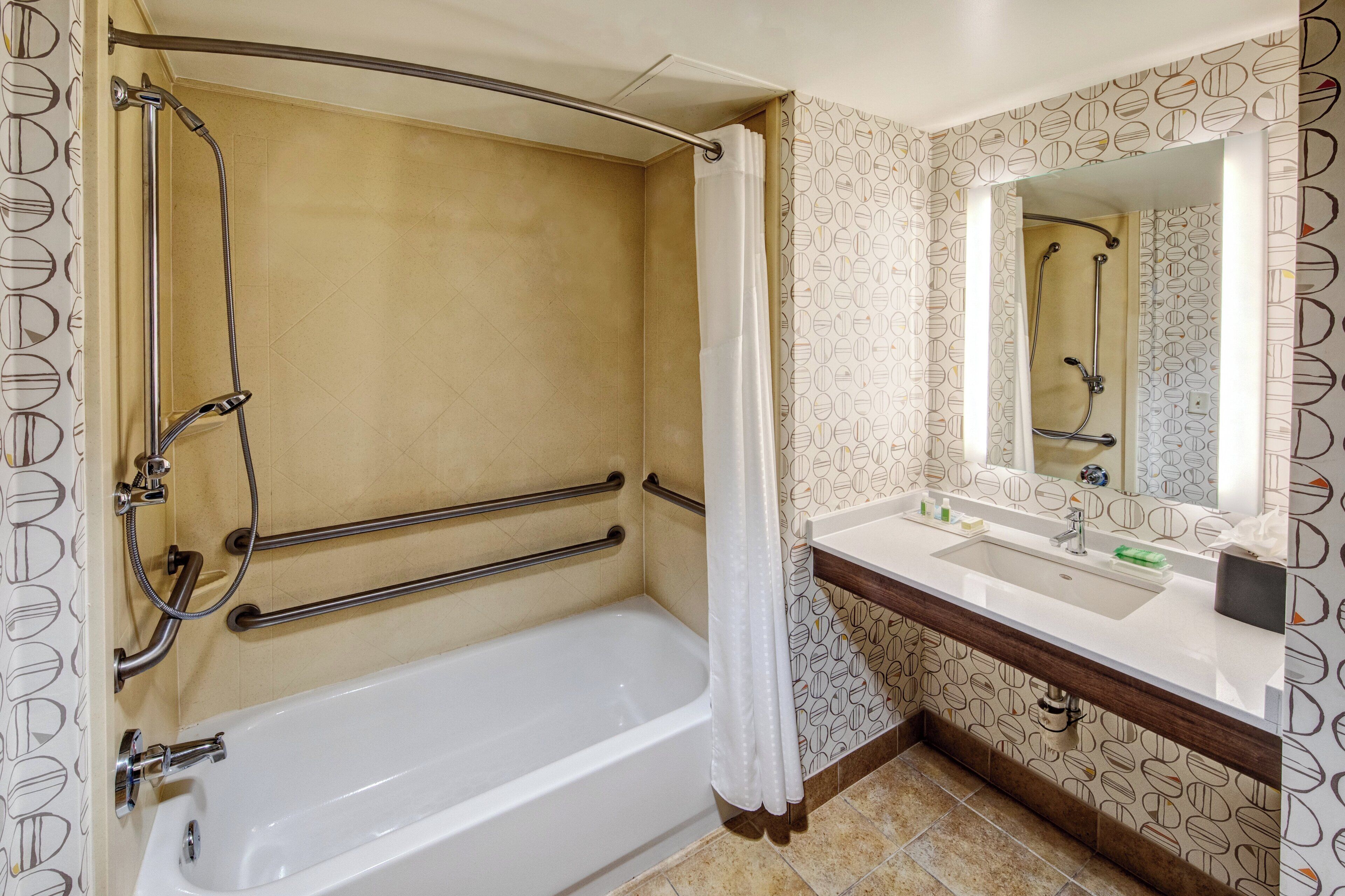 A bathroom with a bathtub, shower, and sink, equipped with grab bars and a mirror.
