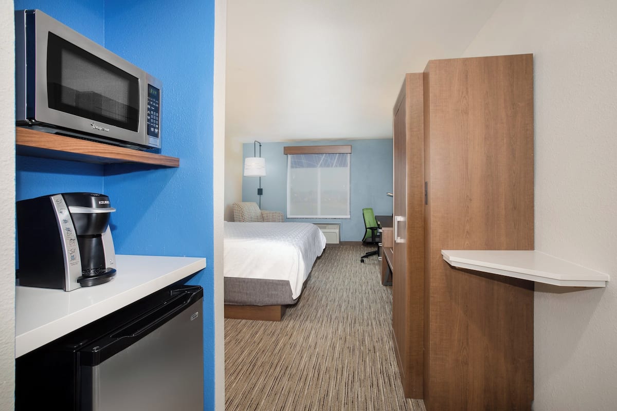 A hotel room with a microwave, coffee maker, bed, bedside table, lamp, and a wooden wardrobe.