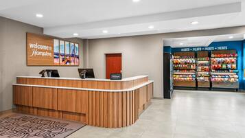 Una mesa de recepción con un cartel que dice 'Welcome to Hampton', una barra de madera, una placa de 'Hilton Honors' y una exhibición de aperitivos.