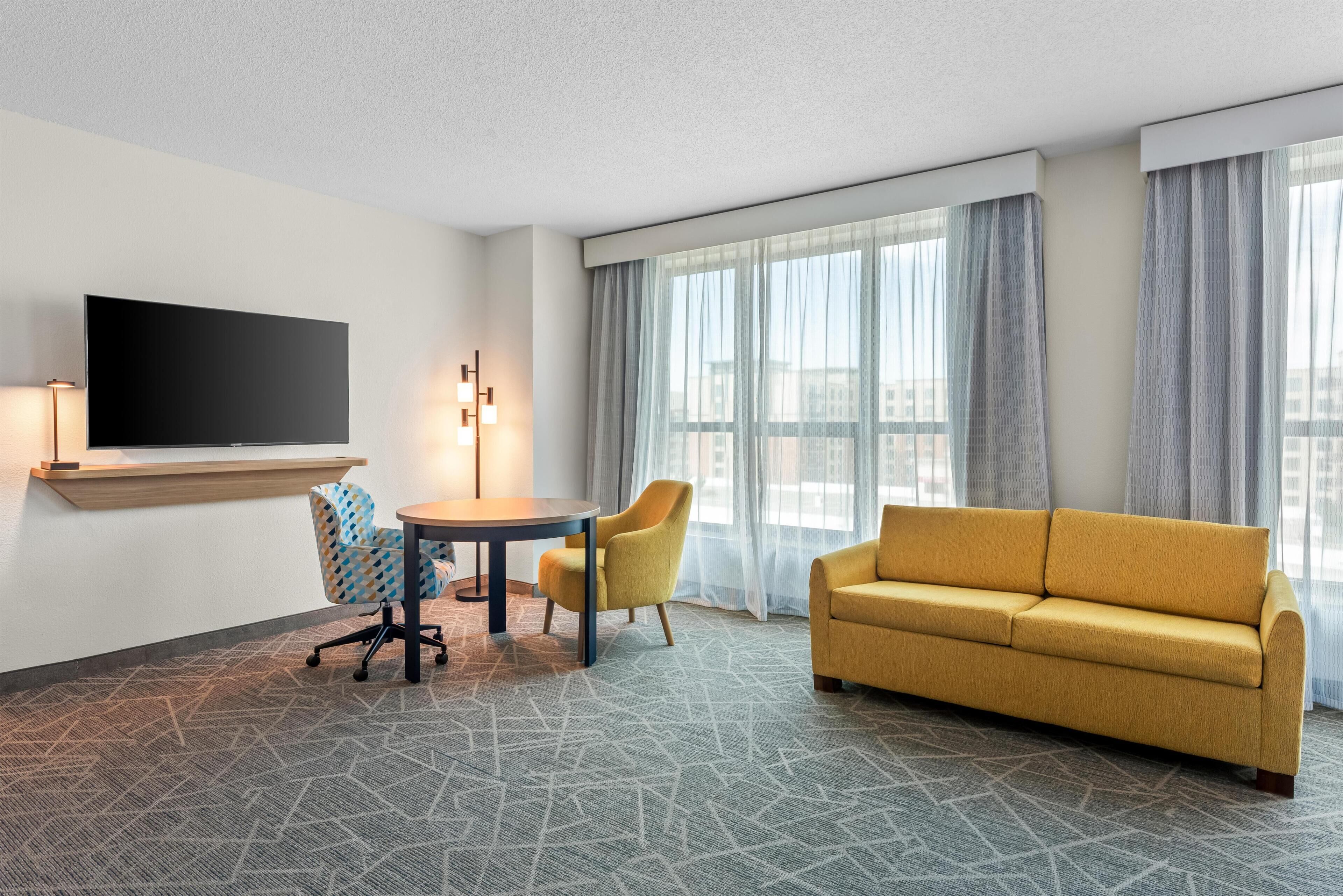 Habitación de hotel con sofá, una mesa con sillas, televisor de pantalla plana en la pared y un ventanal con cortinas.