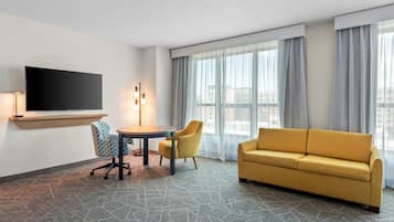 Habitación de hotel con sofá, una mesa con sillas, televisor de pantalla plana en la pared y un ventanal con cortinas.