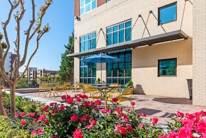 A building with a brick facade, large windows, outdoor seating with yellow chairs, and a blue umbrella.