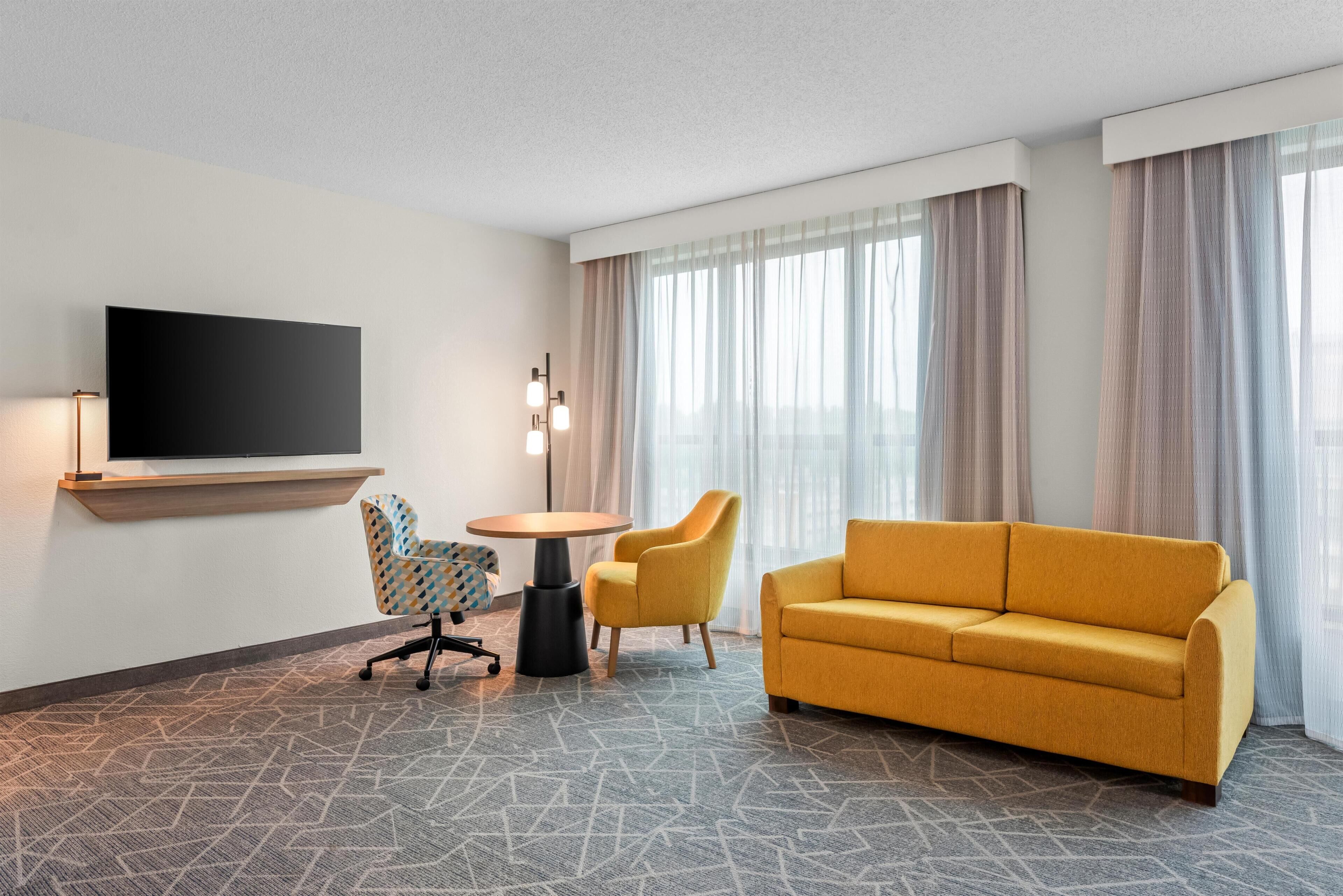 Habitación de hotel con televisor de pantalla plana en la pared, un sofá, dos sillones, una mesita redonda y una lámpara de pie.
