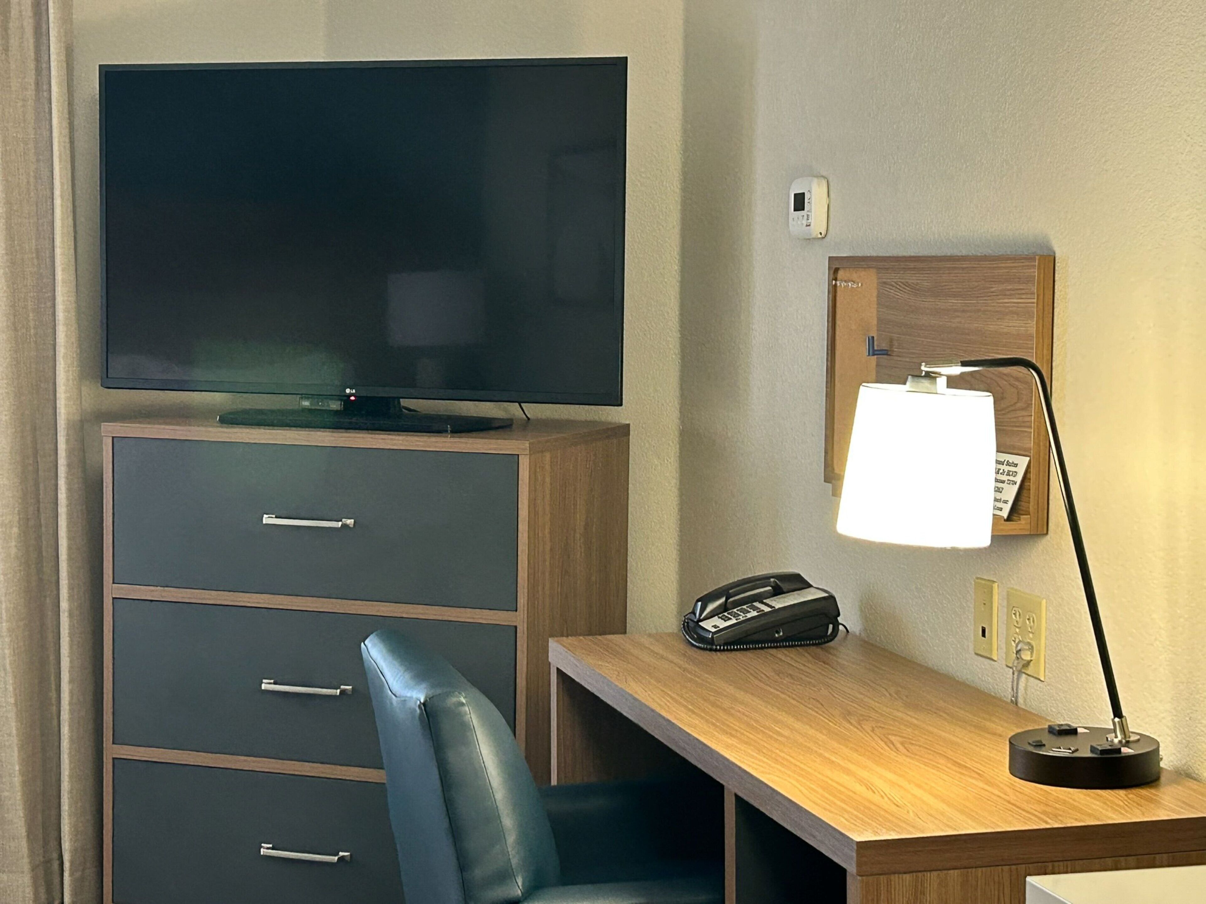 Habitación de hotel con un televisor de pantalla plana sobre un mueble de madera, un escritorio con teléfono y lámpara, y un interruptor de luz en la pared.