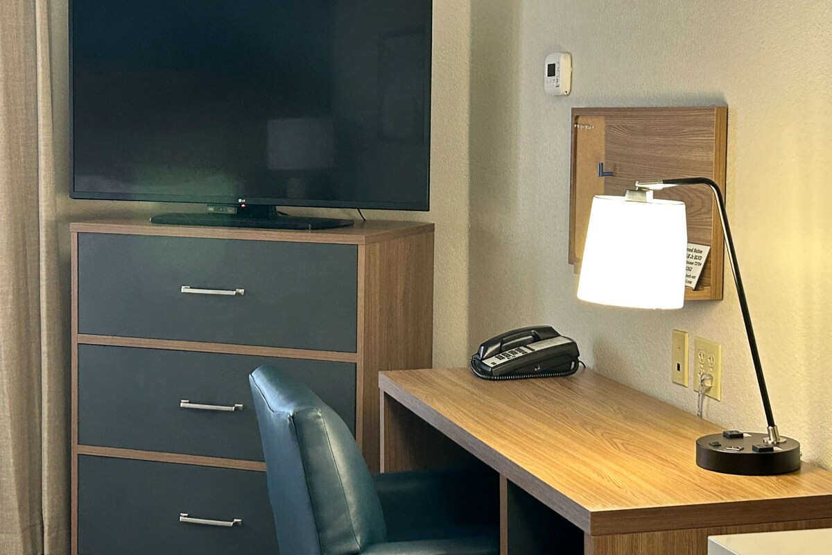 Habitación de hotel con televisión de pantalla plana sobre un mueble de madera, un escritorio con lámpara, teléfono y un tablón de anuncios en la pared.