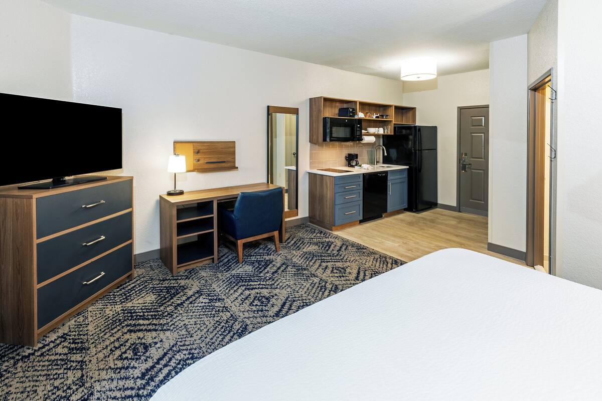 Ett hotellrum med en säng, ett skrivbord med en stol, en tv, en mikrovågsugn, ett kylskåp och ett handfat.