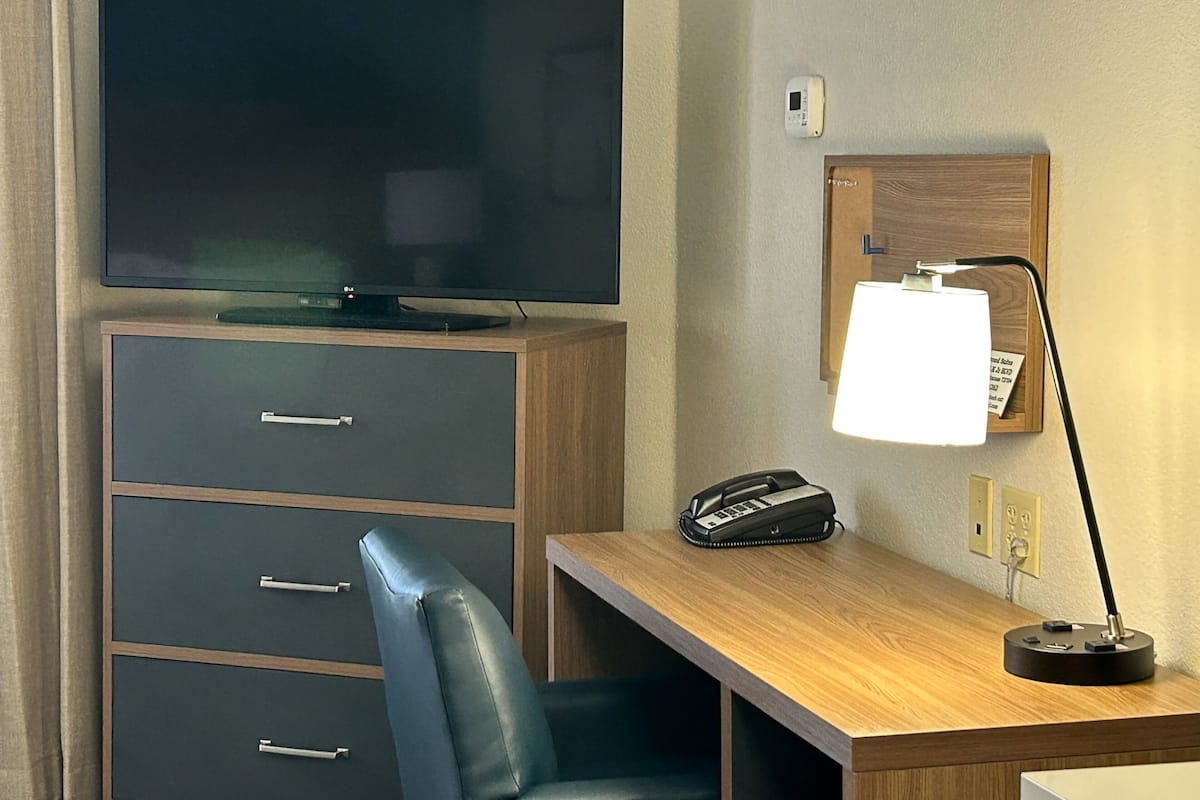 Ett hotellrum med en platt-TV placerad på ett träskåp, ett skrivbord med en lampa, en telefon och en anslagstavla på väggen.