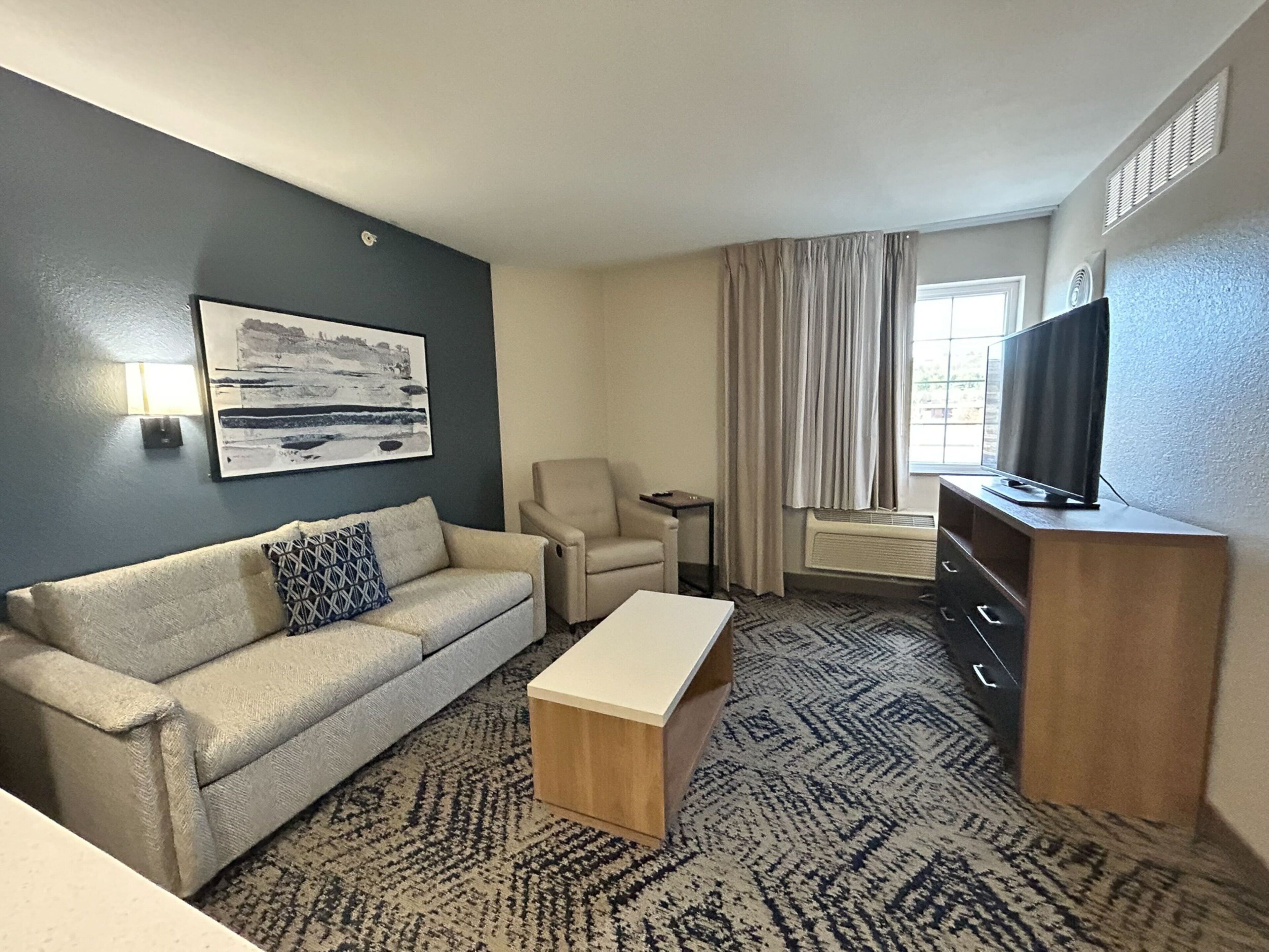 Una habitación de hotel con un sofá color beige, una mesita de madera, un sillón color beige, un televisor sobre un mueble de madera y un ventanal con cortinas.
