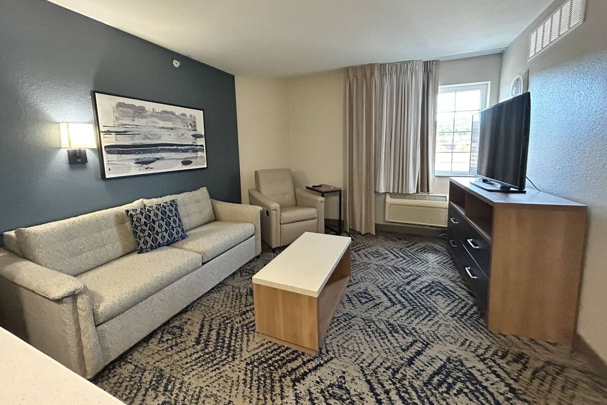 Ett hotellrum med en beige soffa, ett litet träbord, en beige fåtölj, en tv på ett träställ och ett stort fönster med gardiner.