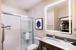 A modern bathroom with a glass shower enclosure, a white toilet, a sink with a marble countertop, and a large mirror with integrated lighting. - Candlewood Suites Fayetteville – University of Arkansas by IHG (Fayetteville)