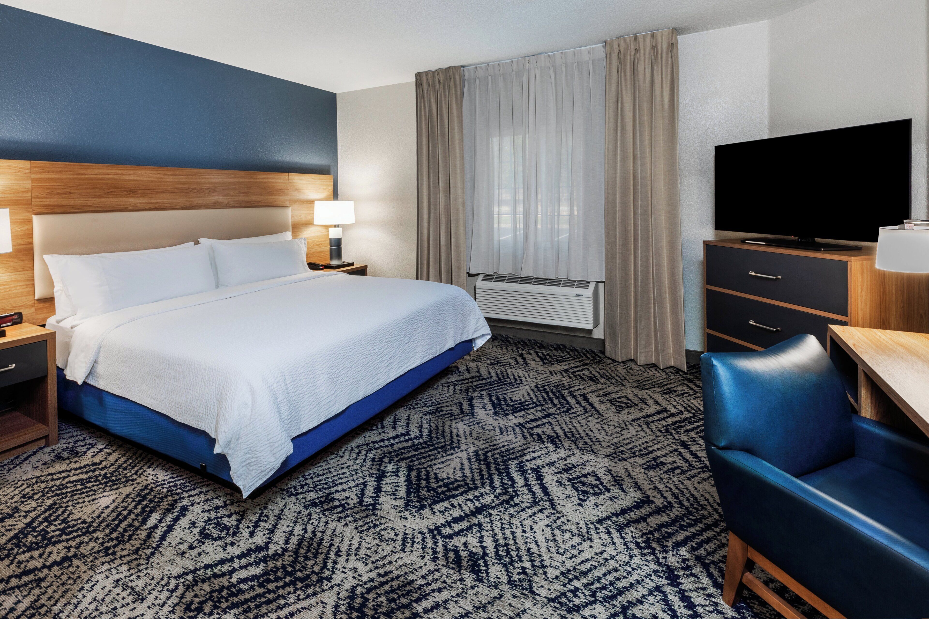 A hotel room with a large bed, a desk with a chair, a flat-screen TV, and a window with curtains.