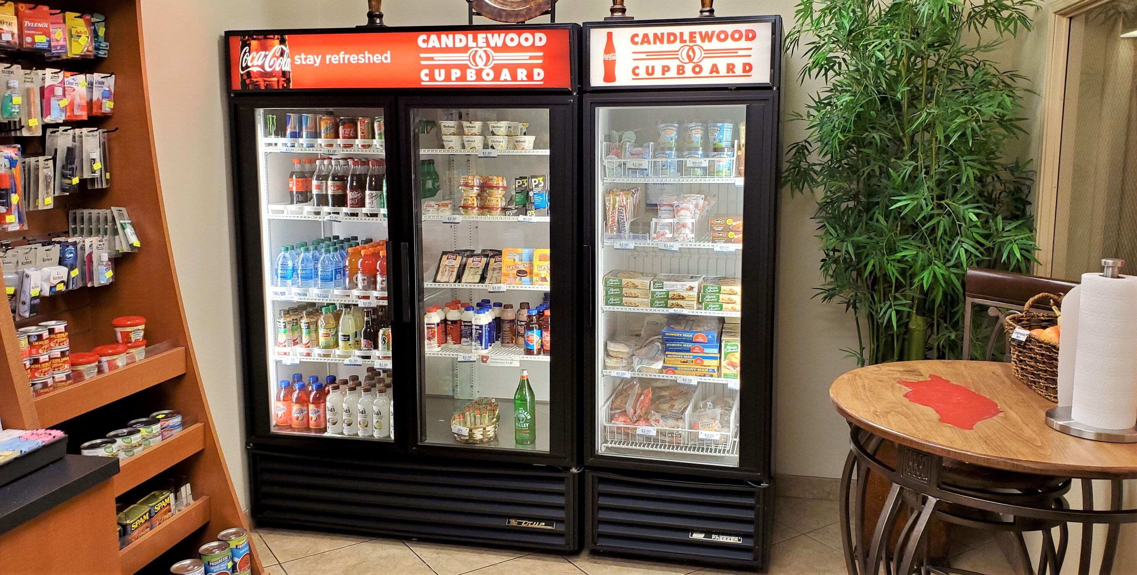 A beverage refrigerator stocked with various drinks and snacks, a Coca-Cola sign, and a Candlewood Cupboard sign.