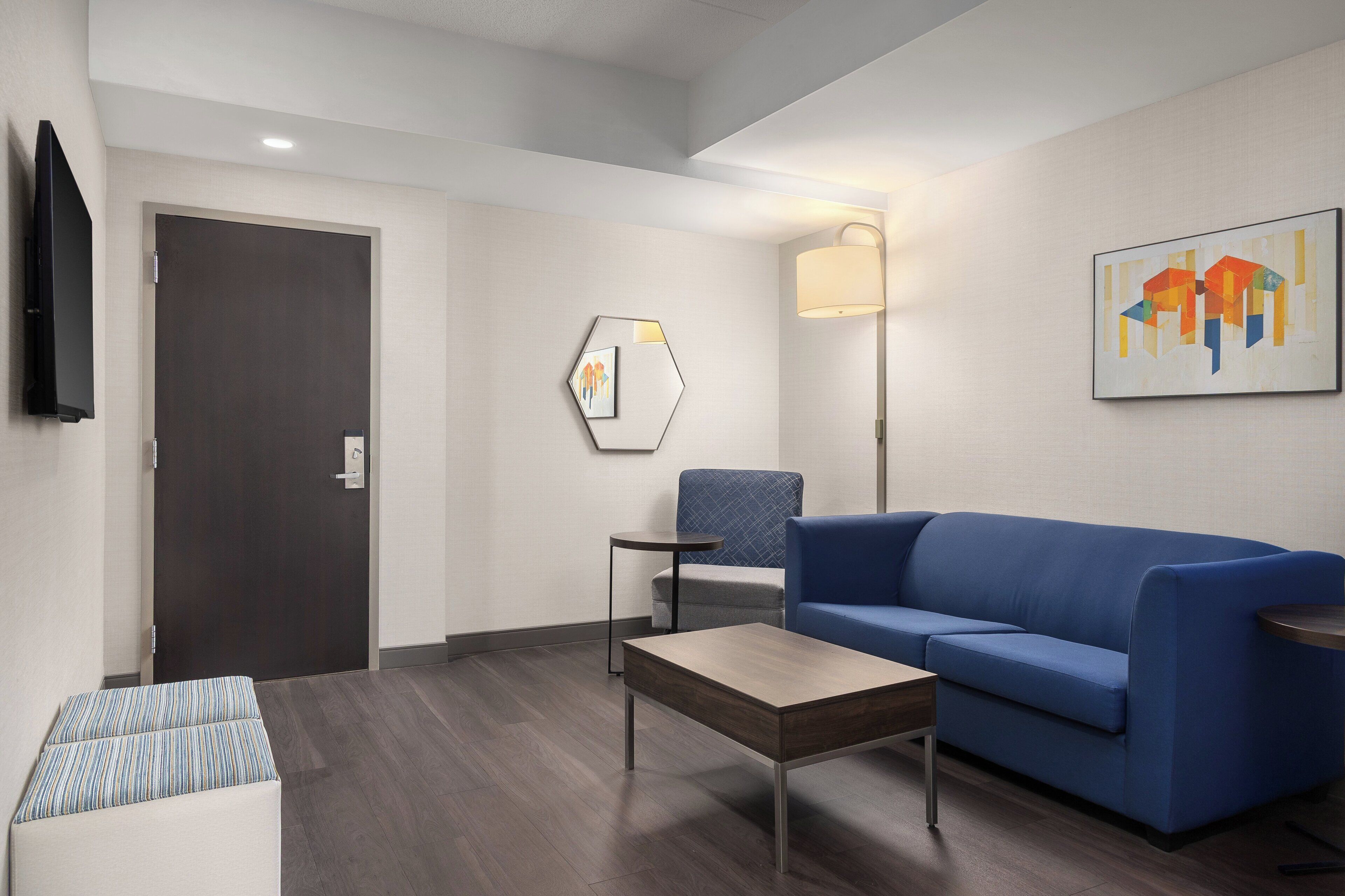 A modern hotel room with a blue sofa, a small table, a lamp, a TV, and a door.