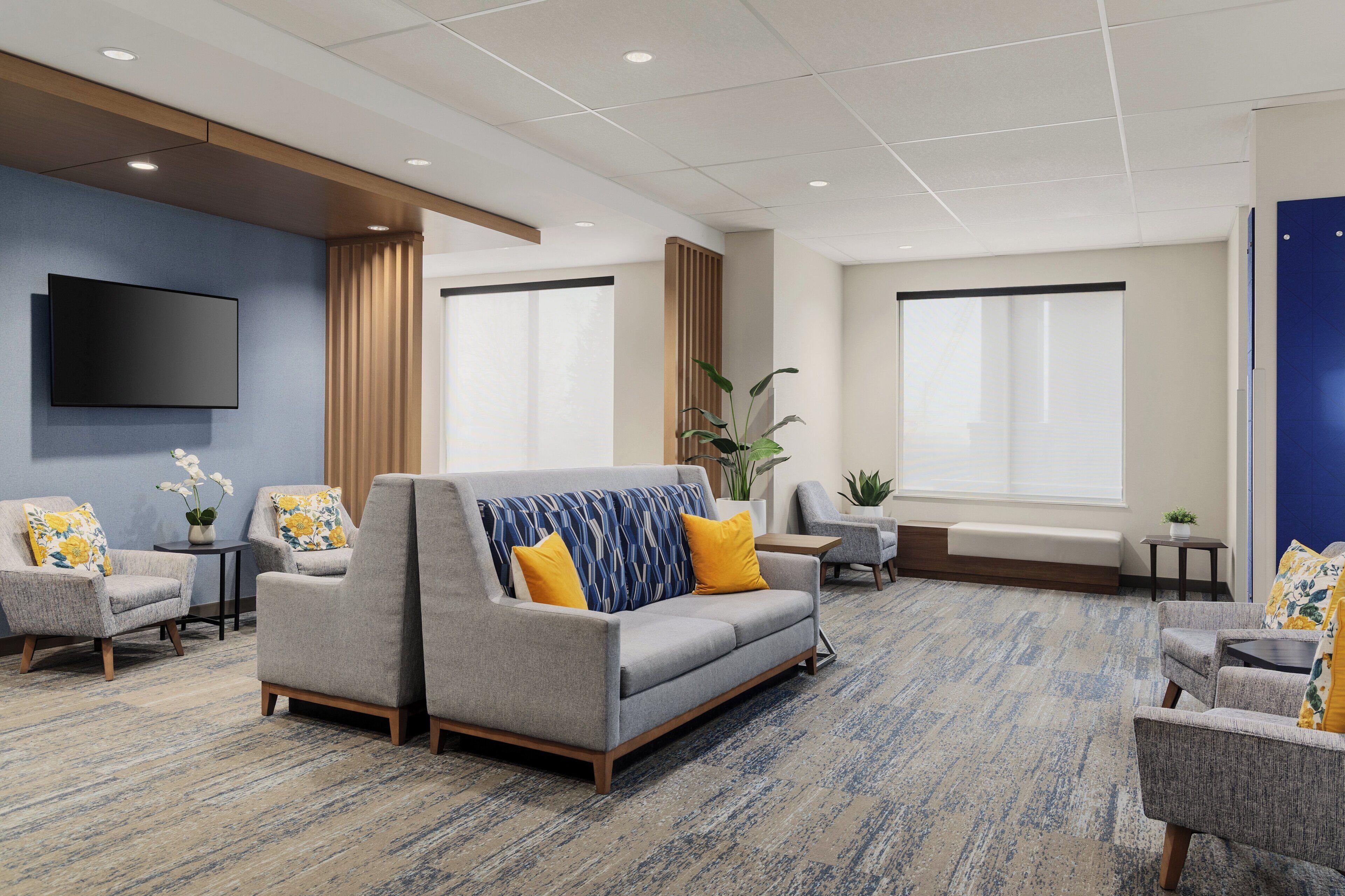 A modern hotel lobby with a grey sofa, armchairs, a flat-screen TV, and large windows.