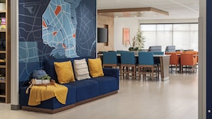 A modern hotel lobby with a blue sofa, yellow cushions, a large abstract wall mural, a dining area with orange chairs, and a flat-screen TV mounted on the wall. - Holiday Inn Express & Suites Vaughan-Southwest by IHG (Vaughan)