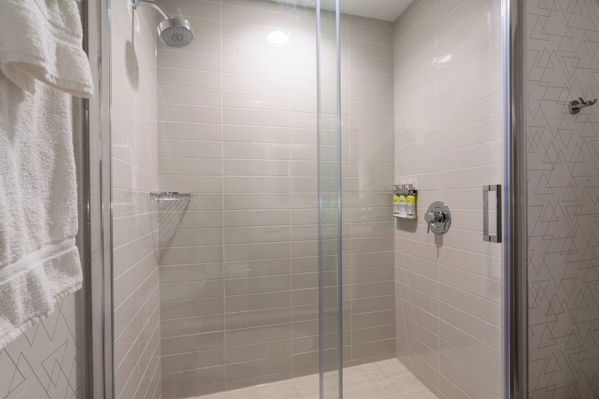 A glass-enclosed shower stall with a handheld showerhead, a towel rack, and a soap dispenser on the wall.