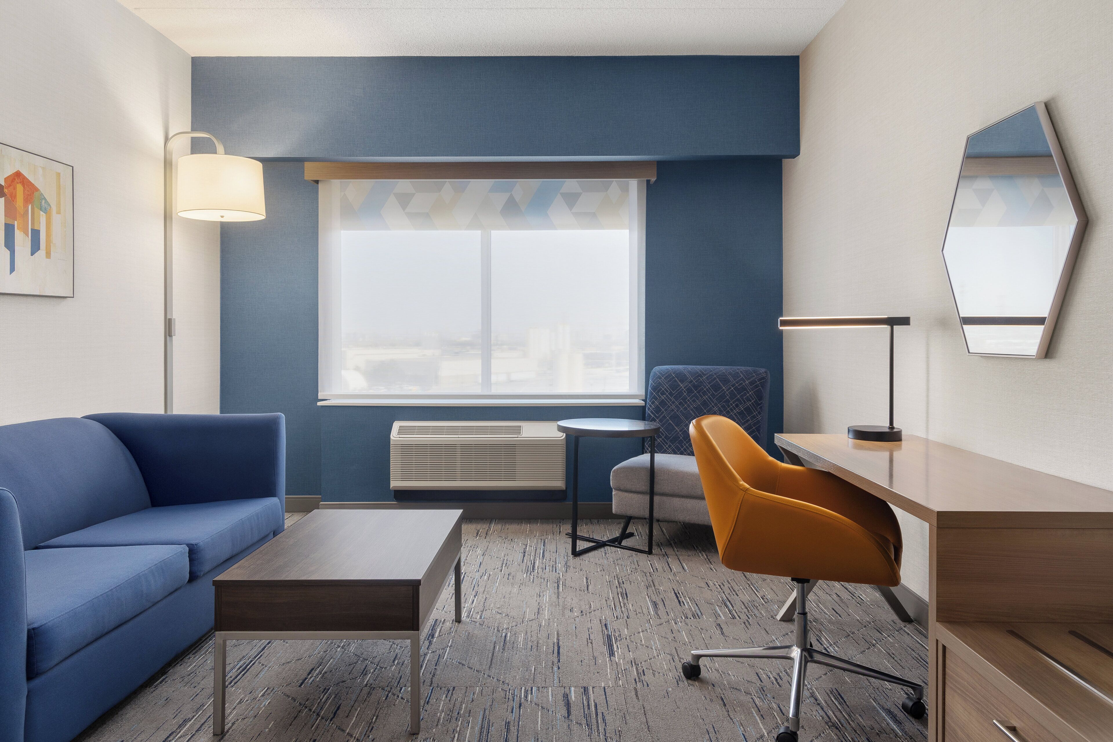 Una habitación de hotel moderna con un sofá azul, un escritorio de madera, una silla amarilla, una lámpara de pie y un ventanal con vistas.