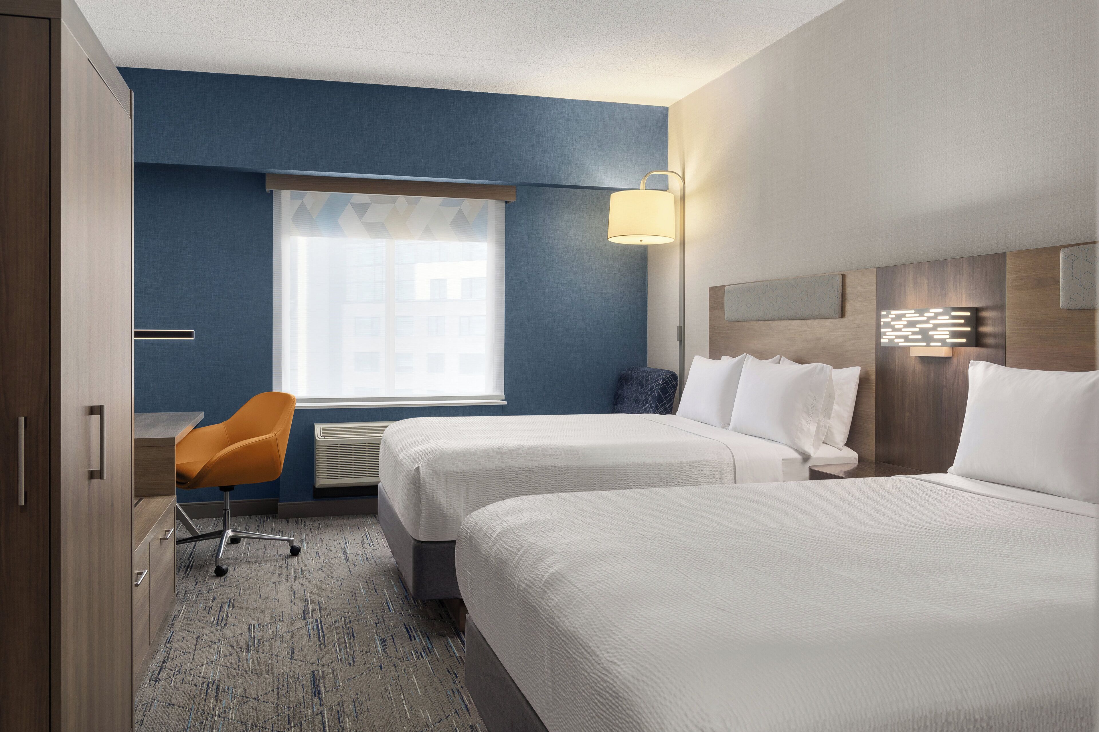 A hotel room with two beds, a desk, an orange chair, a window with blinds, and a wall-mounted lamp.