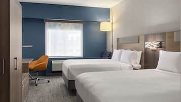 A hotel room with two beds, a desk, an orange chair, a window with blinds, and a wall-mounted lamp.