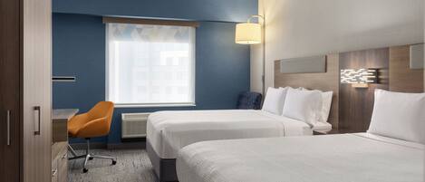 A hotel room with two beds, a desk, an orange chair, a window with blinds, and a wall-mounted lamp.