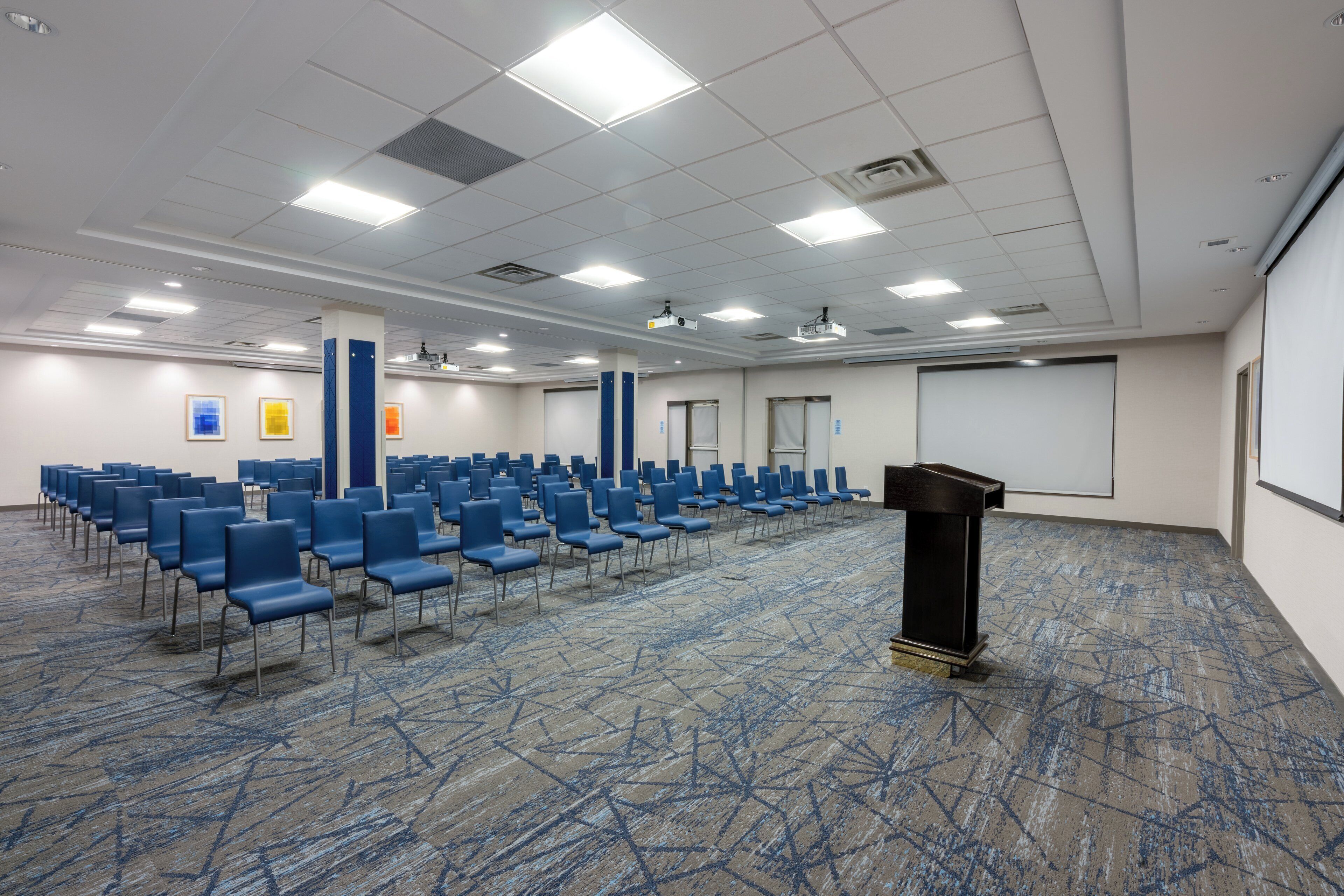 A conference room with rows of blue chairs, a podium, and a projector screen.