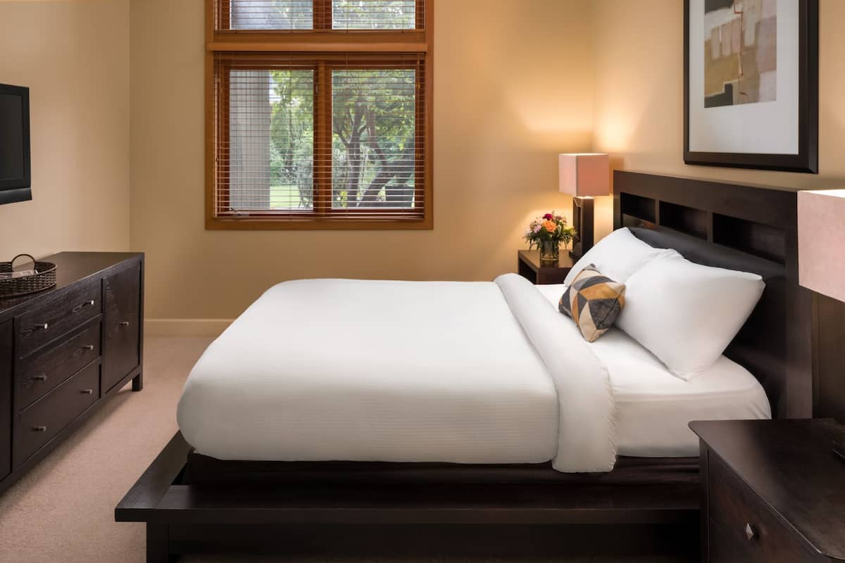 Una cama bien hecha con cabecera de madera, una mesita de noche con lámpara y una ventana con persianas.