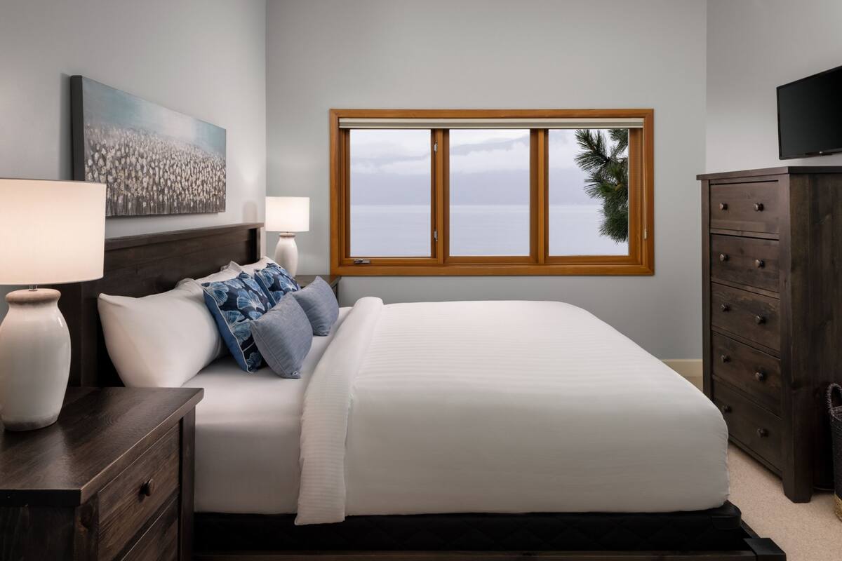 Un dormitorio con una cama, dos mesitas de noche con lámparas, un aparador, un televisor sobre un mueble bajo y una ventana con vistas al océano.