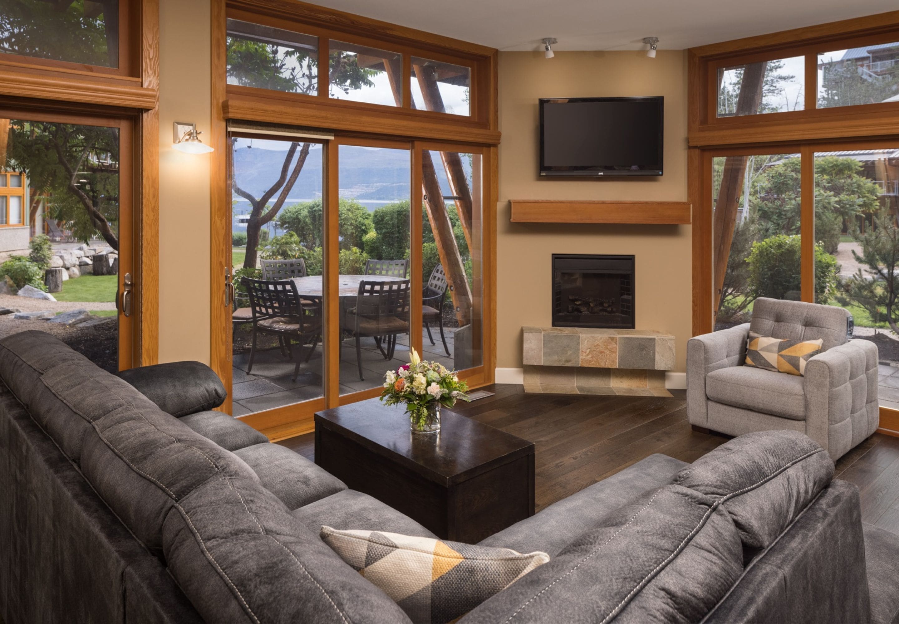 A spacious living room with a large gray sectional sofa, a gray armchair, and a wooden coffee table with a vase of flowers. A flat-screen TV is mounted above a fireplace, and large windows offer a view of the outdoors.