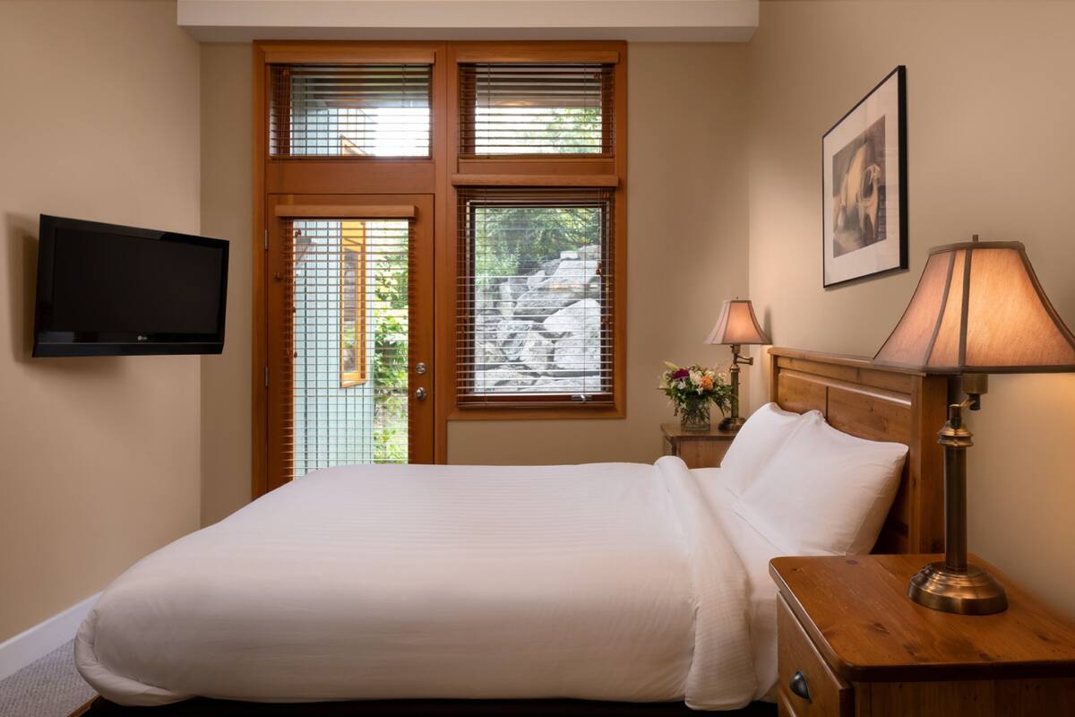 Una cama bien hecha en una habitación de hotel, con un televisor montado en la pared, una lámpara en la mesita de noche y una ventana con persianas.