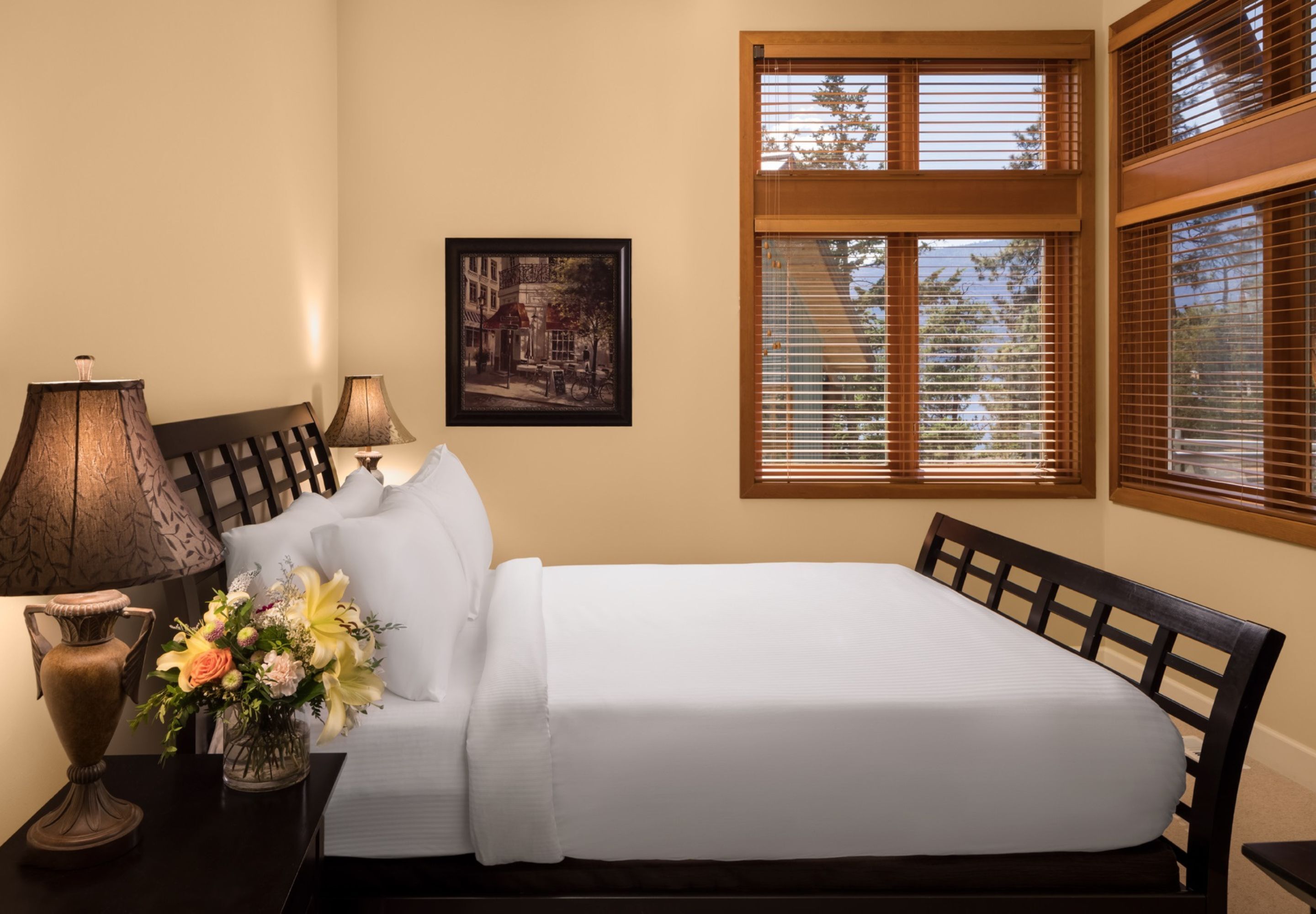 A neatly made bed with white linens, a wooden headboard, and a nightstand with a lamp and vase of flowers.