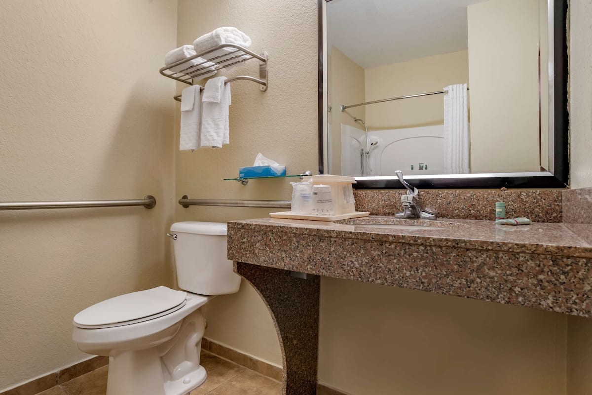 A bathroom with a white toilet, a granite countertop, a towel rack with white towels, a mirror, and a glass shelf with toiletries.