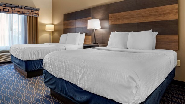 Habitación de hotel con dos camas, un ventanal con cortinas, una lámpara de pie y un panel de madera en la pared.