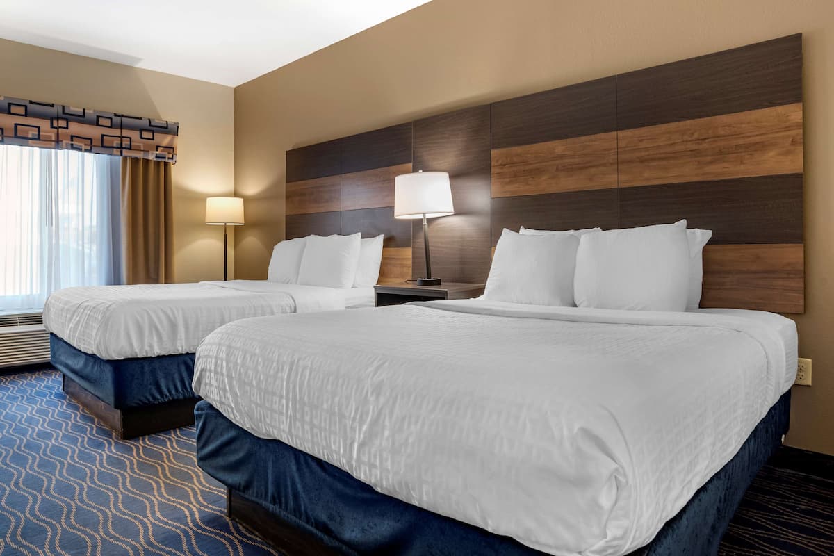A hotel room with two beds, a large window with curtains, a floor lamp, and a wall with a wood panel design.